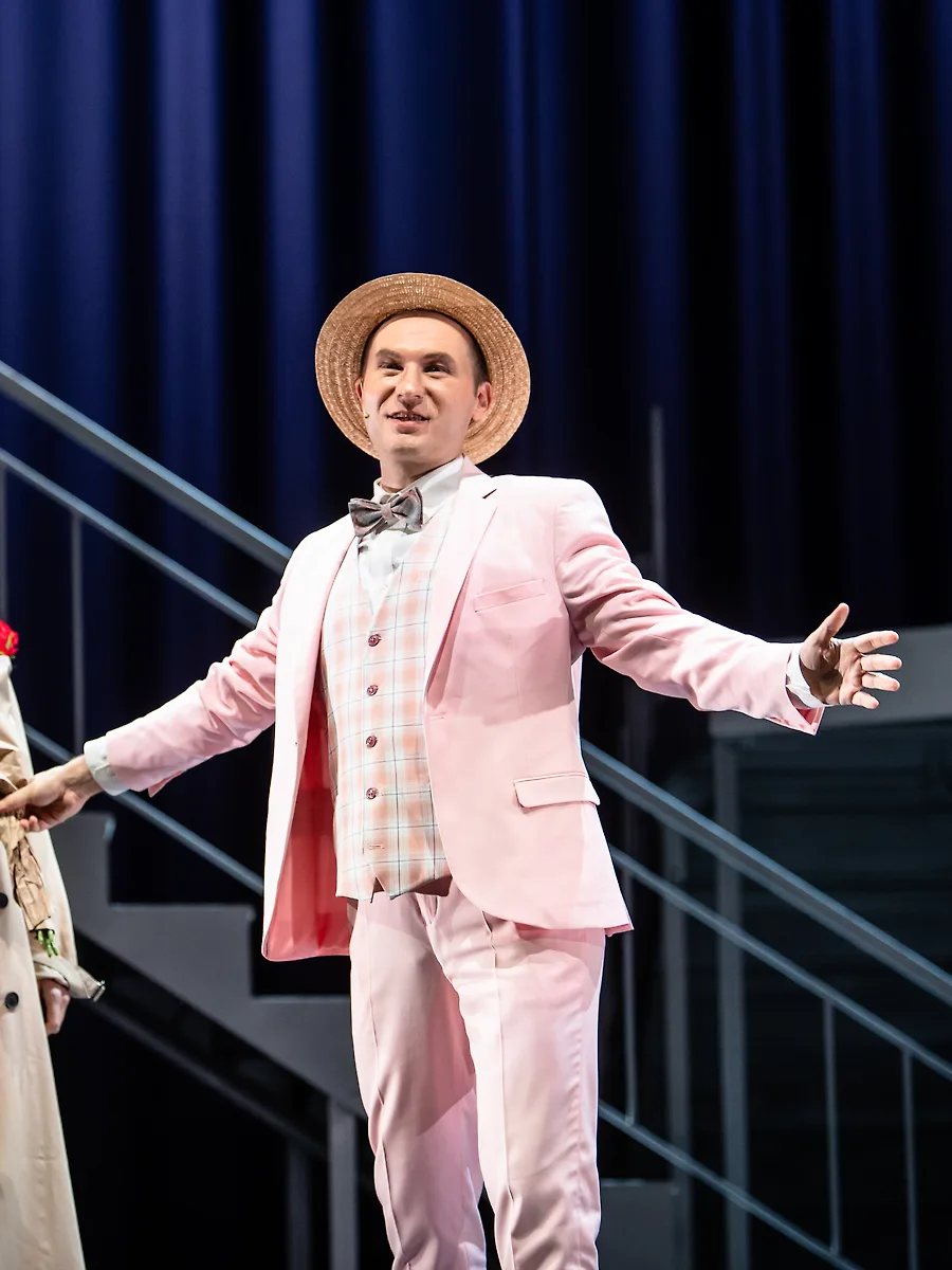Semjon Bulinsky als Freddy in „My Fair Lady“, rosa Anzug und Strohhut, die Arme weit geöffnet, neben ihm eine Frau im hellen Trenchcoat mit roten Rosen, vor tiefblauem Vorhang und einer Treppe