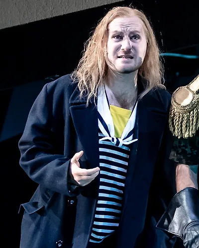 Semjon Bulinsky als Anthony Hope in Sondheims „Sweeney Todd“, lange blonde Haare, Seemannsmantel über gestreiftem Hemd, neben ihm die Schulterklappe des Büttels