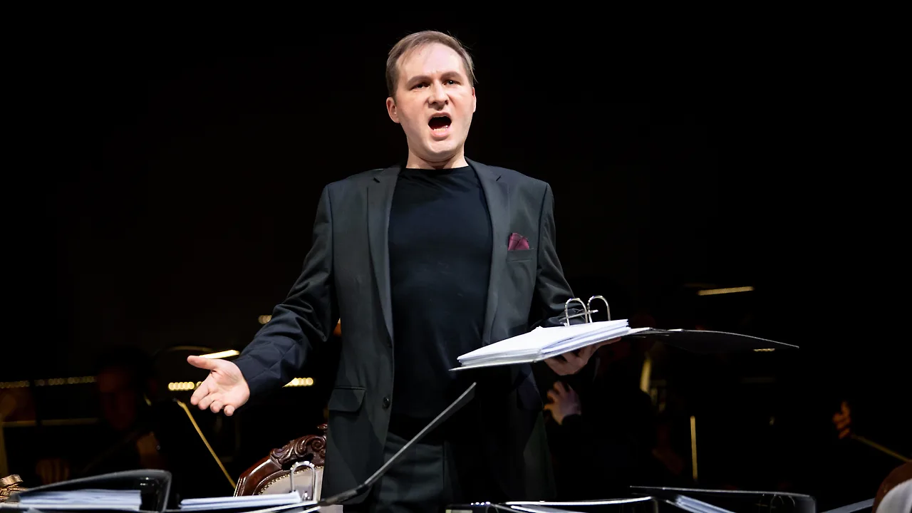 Semjon Bulinsky als Don Ottavio in Mozarts „Don Giovanni“, mitten im Singen mit weit ausgebreiteten Armen, Noten in der Hand, Orchester im Dunkeln dahinter