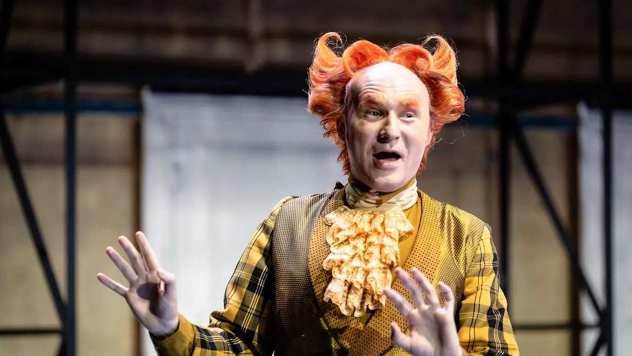 Semjon Bulinsky als Basilio in Mozarts „Le nozze di Figaro“, orange Perücke und gelbes Karokostüm, beide Hände erhoben