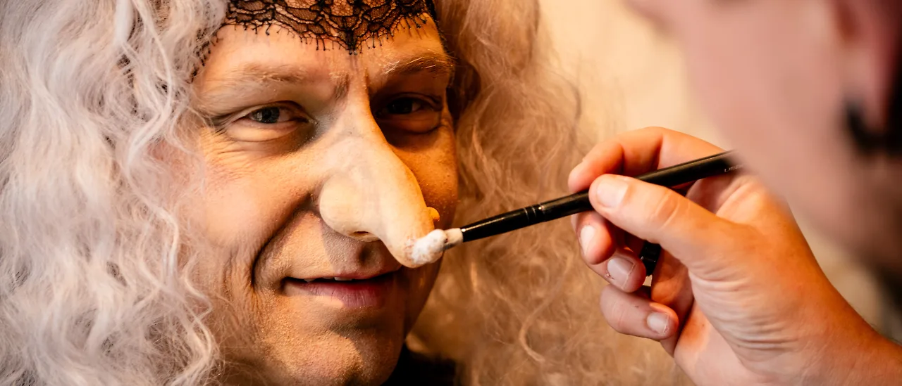 Semjon Bulinsky in der Maske für die Knusperhexe in Humperdincks „Hänsel und Gretel“: graue Lockenperücke, schwarze Spitze über der Stirn, von rechts setzt eine Hand mit dem Pinsel an der angesetzten Nase an