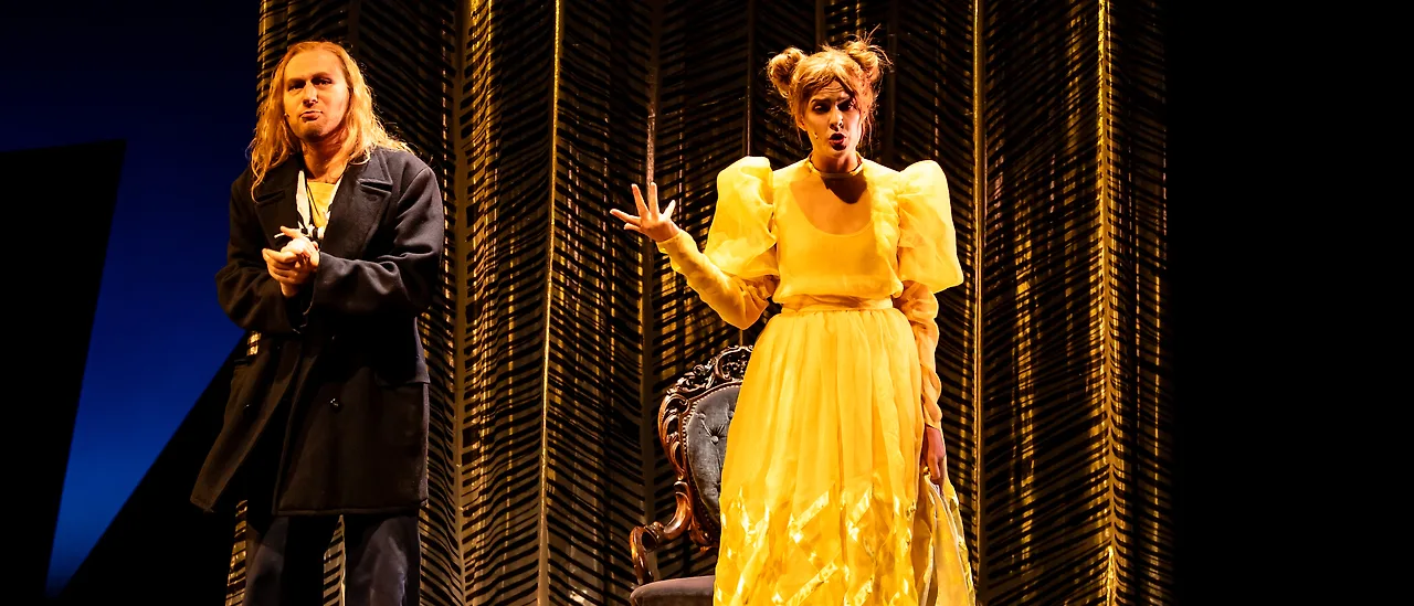Semjon Bulinsky als Anthony Hope in Sondheims „Sweeney Todd“, im dunklen Seemannsmantel vor einem goldenen Vorhang, neben ihm Johanna im gelben Kleid, beide singend