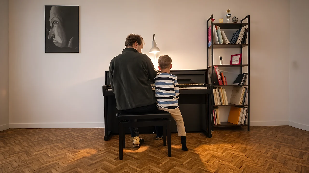 Semjon Bulinsky und ein Schüler sitzen von hinten gesehen nebeneinander an einem schwarzen Klavier in einem hellen Zimmer mit Parkett und Bücherregal