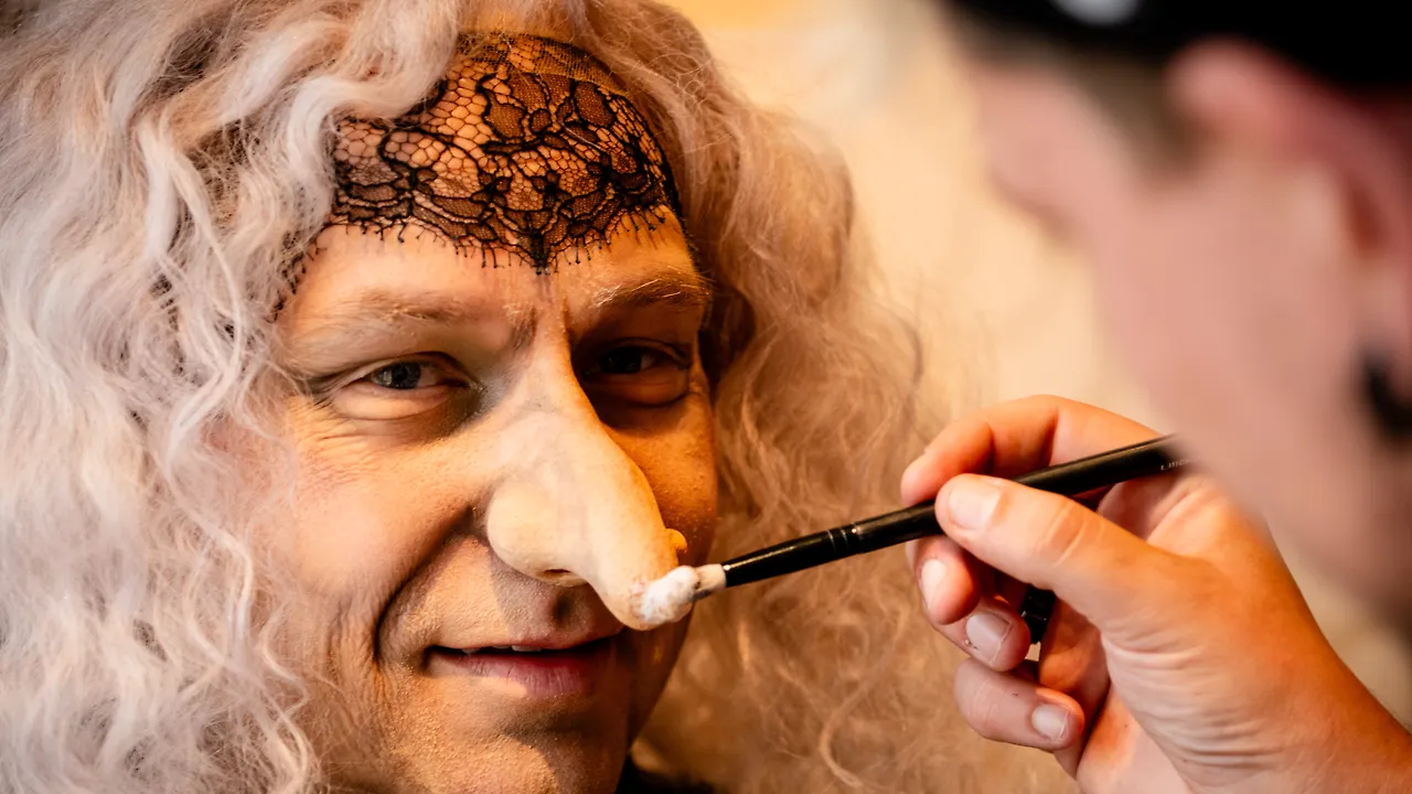 Semjon Bulinsky in der Maske für die Knusperhexe in Humperdincks „Hänsel und Gretel“: graue Lockenperücke, schwarze Spitze über der Stirn, von rechts setzt eine Hand mit dem Pinsel an der angesetzten Nase an