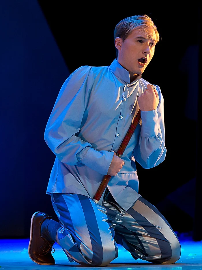 Semjon Bulinsky als Tamino in Mozarts „Zauberflöte“, kniend im hellblauen Kostüm, die Flöte in der Hand, im blauen Bühnenlicht