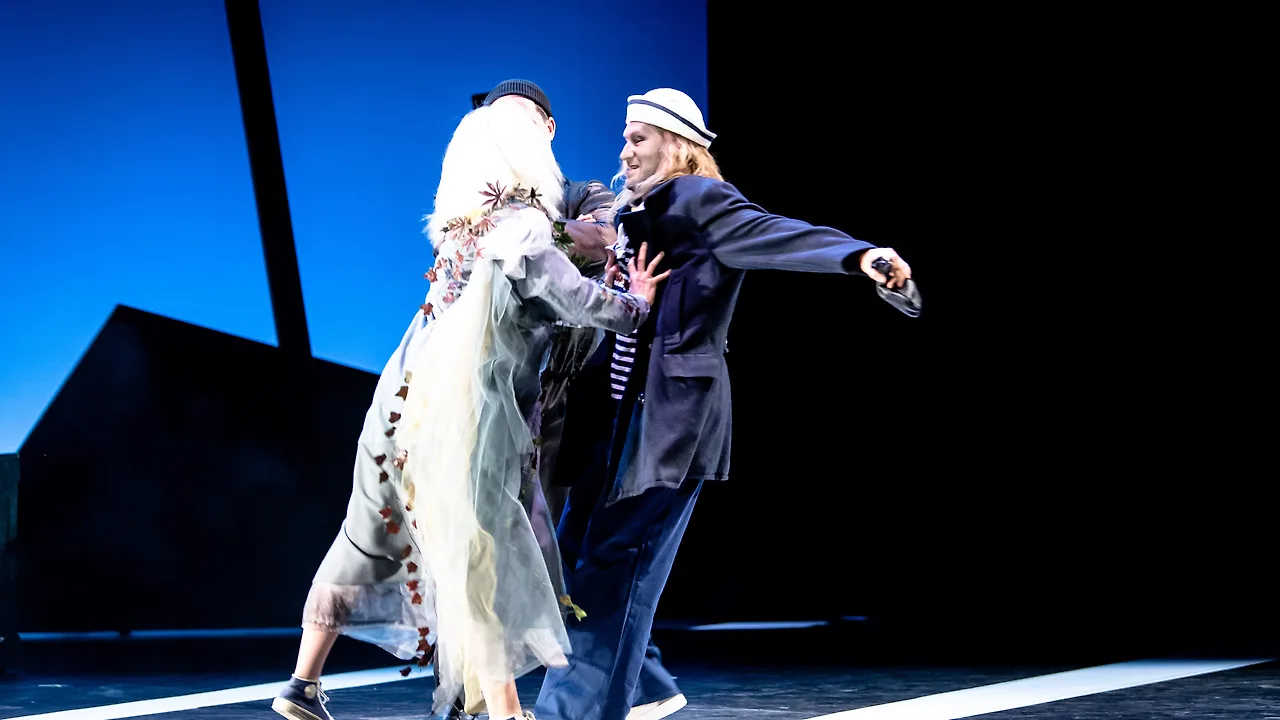 Semjon Bulinsky als Anthony Hope in Sondheims „Sweeney Todd“, im Seemannsmantel mit Matrosenmütze und langen blonden Haaren, lachend, einen Arm weit ausgestreckt; vor ihm eine Frau in weißer Perücke und zerrissenem hellem Kleid, dahinter blauer Himmel und schwarze Silhouetten
